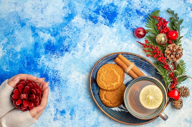 Bovenaanzicht kopje thee citroen schijfjes kaneelstokjes koekjes op schotel rode dennenappel in vrouwelijke hand op blauwe tafel vrije ruimte