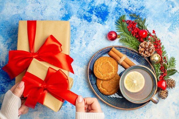 Bovenaanzicht kopje thee kaneelstokjes koekjes op schotel geschenken in vrouwelijke hand xmas boomtak op blauwe tafel