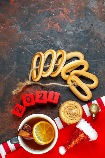 Gratis foto bovenaanzicht kopje thee op smaak gebracht door citroen en kaneel mini kerstmuts op rood tafelkleed houtblokken ovale bagels koekjes op donkerrode tafel