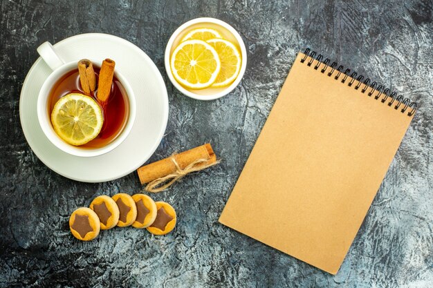 Bovenaanzicht kopje thee op smaak gebracht met citroen en kaneel citroenschijfjes in kleine schotel koekjes kaneelstokjes notitieboekje op donkere tafel