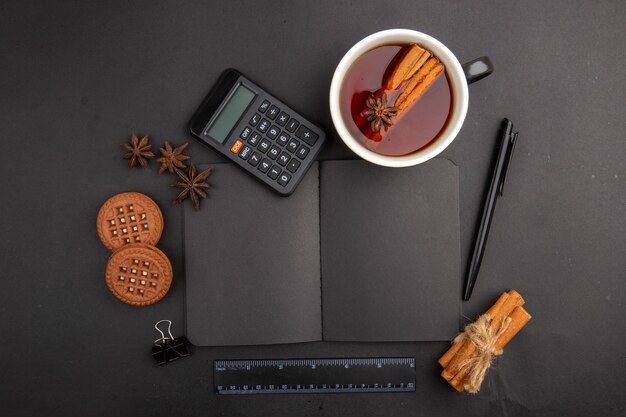 Bovenaanzicht kopje thee op smaak gebracht met kaneel en anijs rekenmachine notitieblok koekjes pen liniaal op donkere tafel