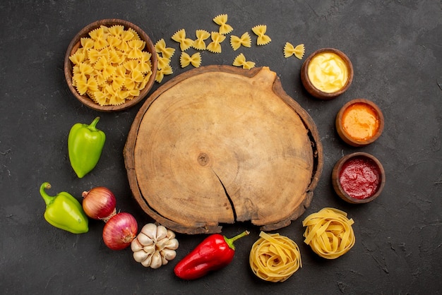 Bovenaanzicht kruiden pasta in kom drie soorten saus knoflook ui rode en groene paprika naast de houten snijplank