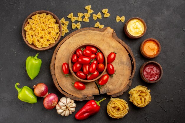 Bovenaanzicht kruiden pasta in kom drie soorten saus knoflook ui rode en groene paprika naast de tomaten op de houten snijplank