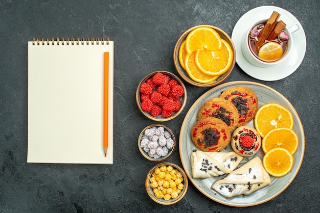 Gratis foto bovenaanzicht lekkere koekjes met gebak, sinaasappelschijfjes, noten en kopje thee op de donkere achtergrond, theenootkoekje, zoete cake