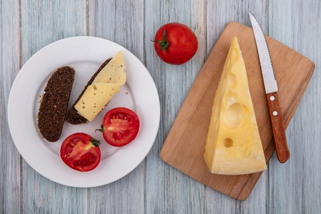 Bovenaanzicht maasdam kaas met een mes op een stand met tomaten en sneetjes zwart brood op een bord op een grijze achtergrond