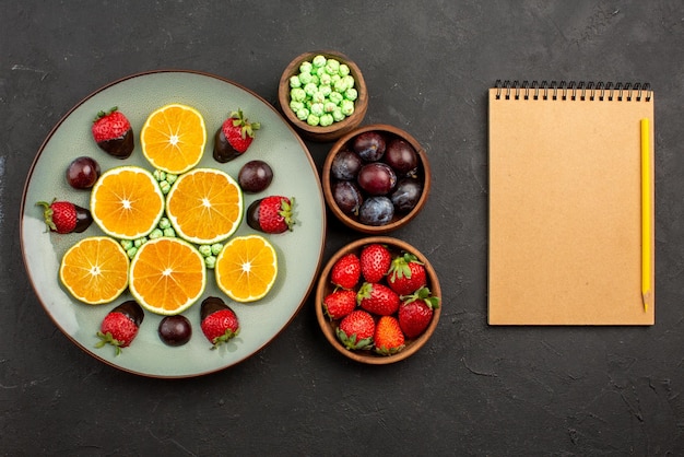 Bovenaanzicht met chocolade bedekte aardbei gehakte sinaasappel met chocolade bedekte aardbei en groene snoepjes en kommen met verschillende soorten fruit, bessen en snoep naast het notitieboekje en het potlood