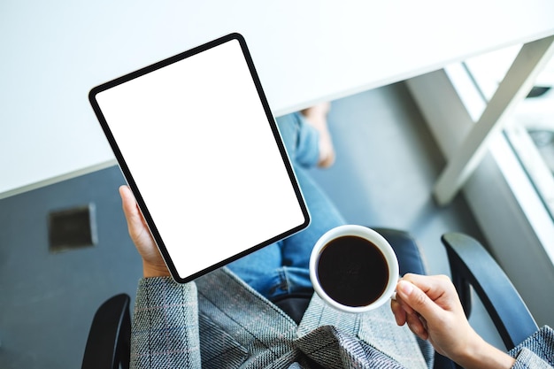 Bovenaanzicht mockup-afbeelding van een zakenvrouw die een digitale tablet vasthoudt met een leeg wit bureaubladscherm terwijl ze koffie drinkt op kantoor