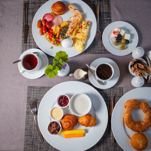 Bovenaanzicht ontbijt eieren, olijven, omelet, croissantjes in borden en kopje thee op tafel