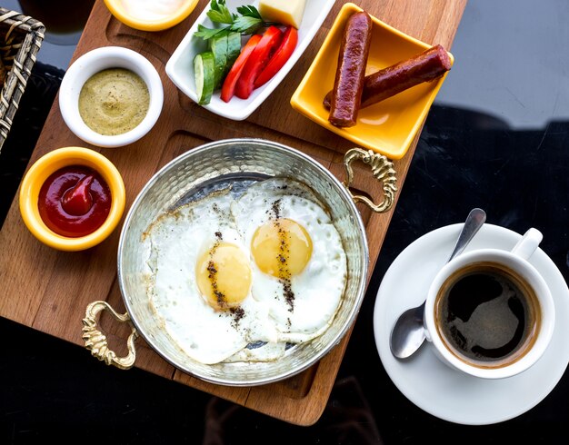 Bovenaanzicht ontbijt gebakken eieren in een pan met worstjes en groenten met sauzen en een kopje koffie