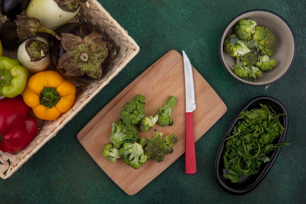 Bovenaanzicht paprika met aubergines in een mand met broccoli op een snijplank met een mes op een groene achtergrond