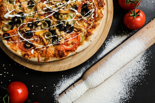 Bovenaanzicht pizza op stand met tomaten olijven deegroller en meel op zwarte tafel