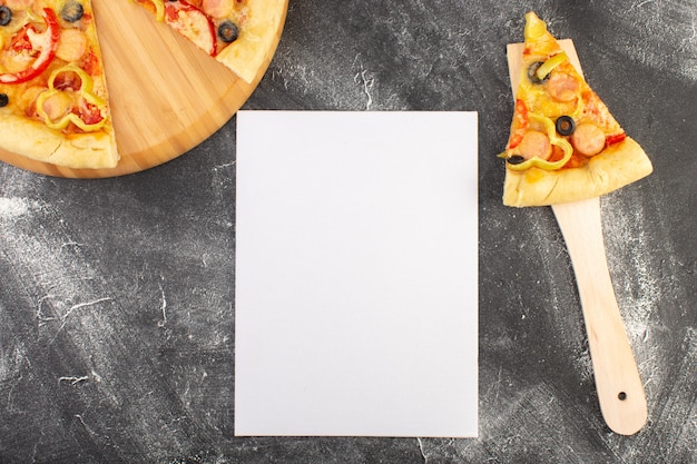 Bovenaanzicht pizzaplak met zwarte olijven tomaten en worst op de houten lepel in de buurt van leeg papier blanco op het grijze bureau eten italiaanse deeg pizzamaaltijd