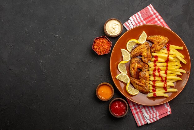 Bovenaanzicht plaat van fastfood kippenvleugels frietjes met citroen en ketchup en kommen met sauzen en kruiden op roze-wit geruit tafelkleed aan de rechterkant van zwarte tafel