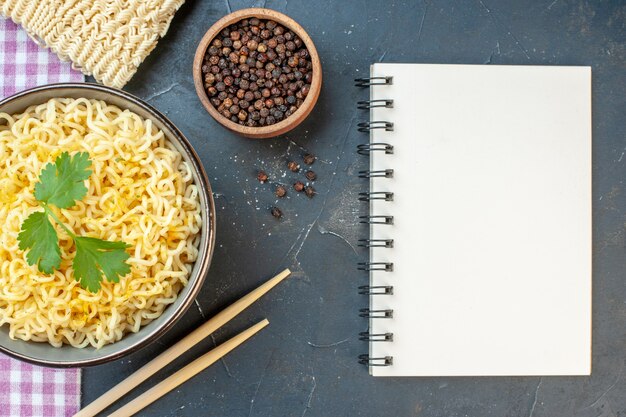 Bovenaanzicht ramen noodles met koriander in kom eetstokjes zwarte peper in kleine kom rauwe noedels notitieblok op donkere tafel