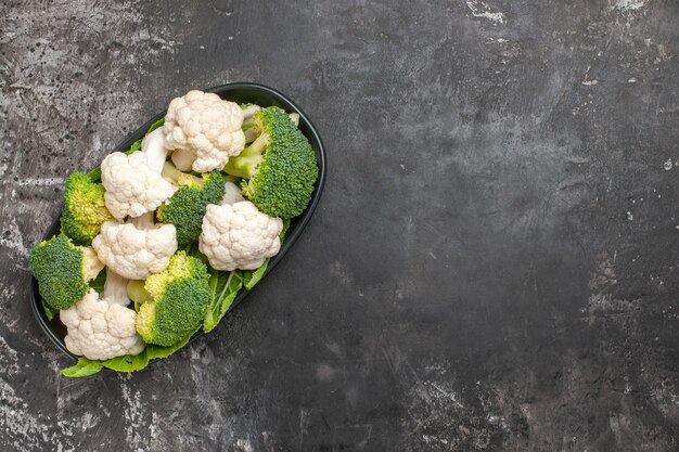 Bovenaanzicht rauwe broccoli en bloemkool op zwarte ovale plaat op donkere ondergrond met kopie ruimte