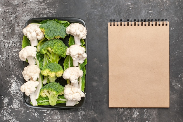 Bovenaanzicht rauwe broccoli en bloemkool op zwarte rechthoekige plaat een notitieboekje op donkere ondergrond