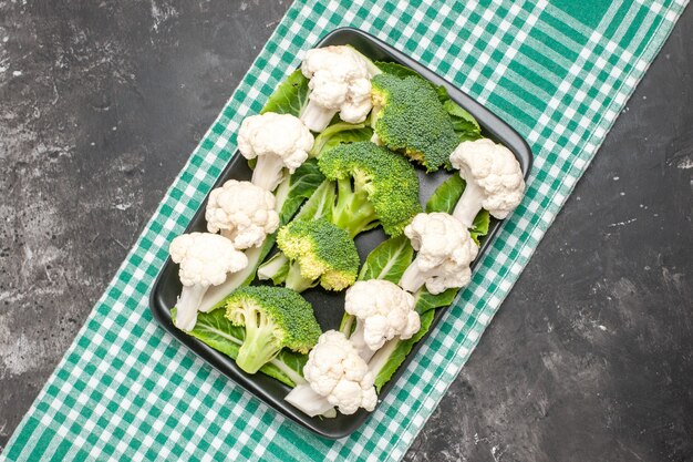 Bovenaanzicht rauwe broccoli en bloemkool op zwarte rechthoekige plaat op groen en wit geruit tafelkleed op donkere ondergrond