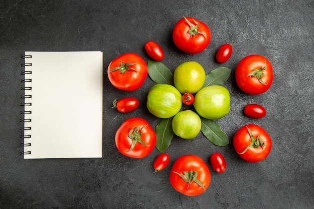 Bovenaanzicht rode en groene tomaten laurierblaadjes rond een kerstomaat en een notitieboekje op donkere grond