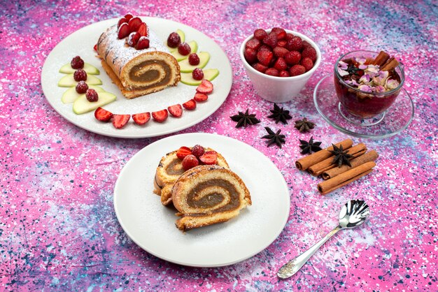 Bovenaanzicht roltaart in bord met appels en aardbeien, samen met kaneel en thee op het helderpaarse bureaukoekkoekje met zoet fruit