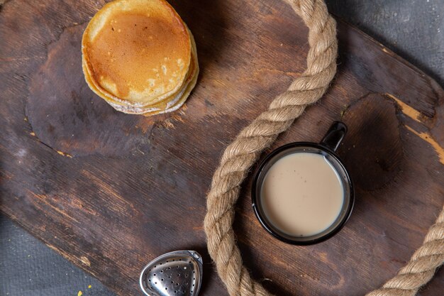 Bovenaanzicht ronde pannenkoeken met zwarte kop melk op het houten bureau met touwen op de grijze achtergrond voedsel maaltijd ontbijt zoet