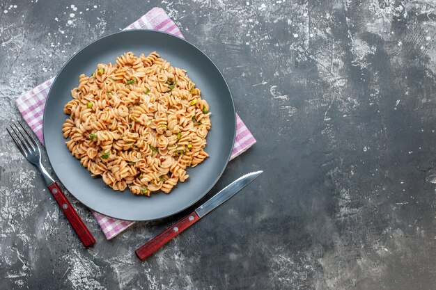 Bovenaanzicht rotini pasta op ronde plaat op roze wit geruite servetvork en mes op grijze tafel vrije ruimte