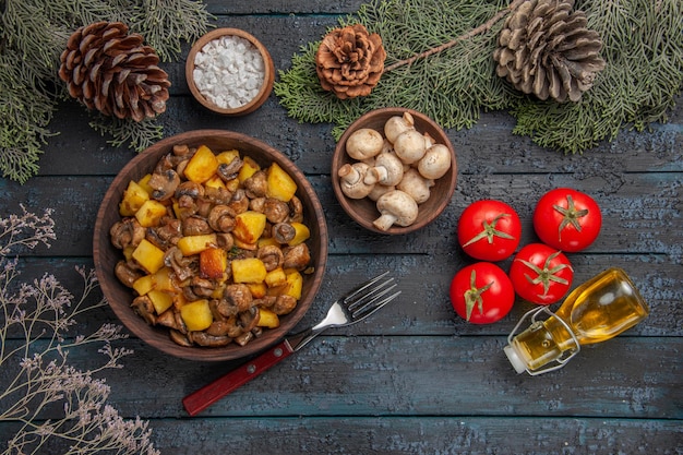 Bovenaanzicht schotel en takken plaat van champignons en aardappelen op de grijze tafel onder de vuren takken met kegels champignons en zout naast de vork tomaten en olie