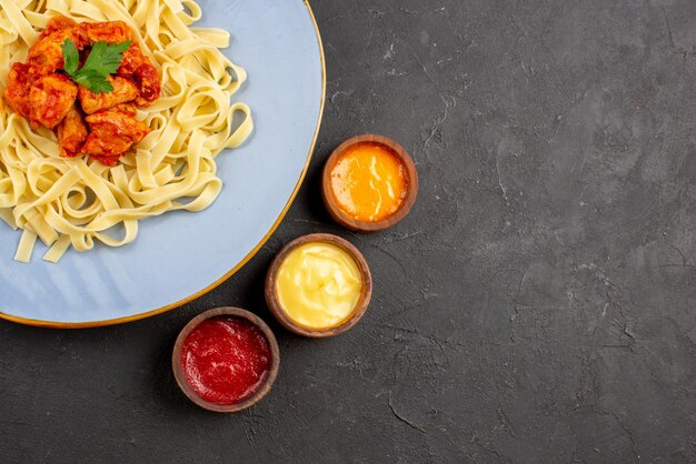 Bovenaanzicht schotel met sauzen pasta met jus en vlees in de blauwe plaat naast de drie soorten sauzen op de donkere achtergrond