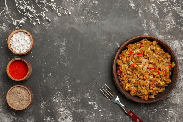 Bovenaanzicht schotel van sperziebonen drie kommen kruiden naast het bord sperziebonen met tomaten en vork op de zwarte tafel