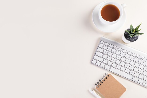 Bovenaanzicht shot van een toetsenbord, notitieblok, pen, een kop warme koffie en een plant op witte tafel