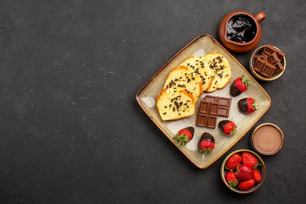 Bovenaanzicht smakelijke cakecake met aardbeien en chocolade tussen kommen chocoladeroomaardbeien en chocolade aan de rechterkant van de zwarte tafel