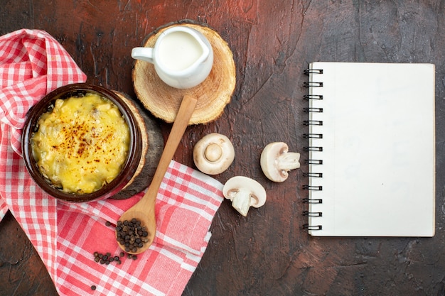 Bovenaanzicht smakelijke julienne in kom rauwe champignons melkkom op houten bord zwarte peper in houten lepel notitieboekje op donkerrode tafel