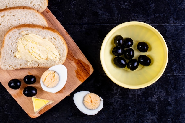 Bovenaanzicht sneetjes brood en boter aan boord met olijven en gekookt ei op zwart