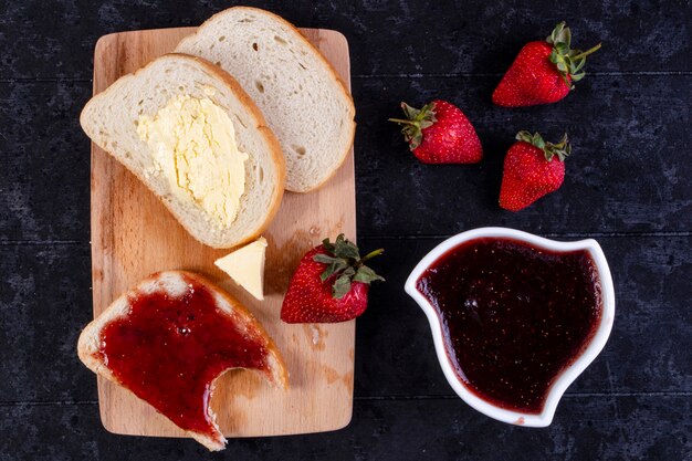 Bovenaanzicht sneetjes brood en boter met een sneetje brood met jam aan boord met aardbeien en jam in een schotel