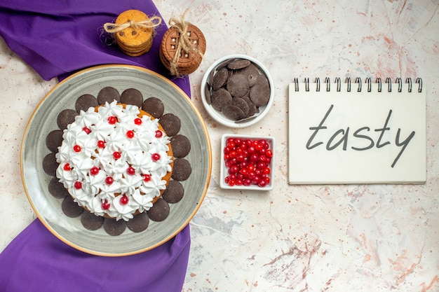 Bovenaanzicht taart met banketbakkersroom op ovale plaat paarse sjaal koekjes vastgebonden met touw chocolade en bessen in kommen lekker geschreven op notitieboekje