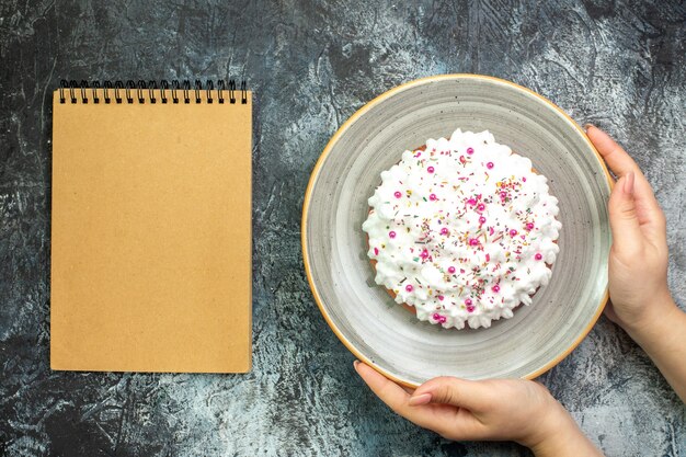 Bovenaanzicht taart met witte banketbakkersroom op grijze ronde schotel in vrouwelijke hand notebook op grijze tafel