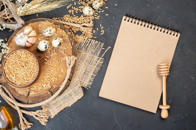 Bovenaanzicht tarwekorrels in kom knoflook op natuurlijk houten bord kwarteleitjes notitieboekje honingstok op tafel