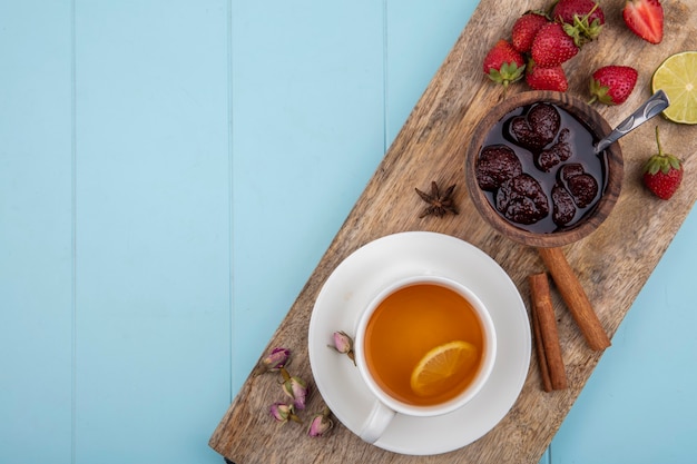 Bovenaanzicht van aardbeienjam op een houten keukenbord met een kopje thee met kaneelstokjes op een blauwe achtergrond met kopie ruimte