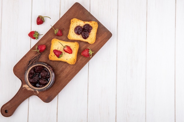 Bovenaanzicht van aardbeienjam op een houten keukenbord met geroosterd brood op een witte houten achtergrond met kopie ruimte