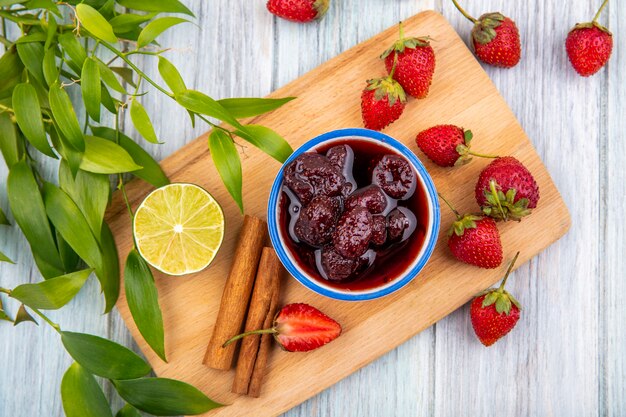 Bovenaanzicht van aardbeienjam op een kom op een houten keukenbord met verse aardbeien met limoen met bladeren op een grijze houten achtergrond