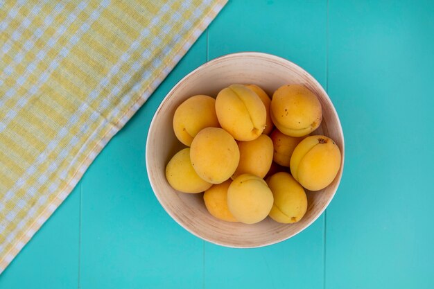 Bovenaanzicht van abrikozen in een kom met een gele geruite handdoek op een blauwe ondergrond