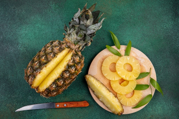 Bovenaanzicht van ananas met een stuk uitgesneden van hele vruchten en ananas segmenten op snijplank met mes op groen oppervlak