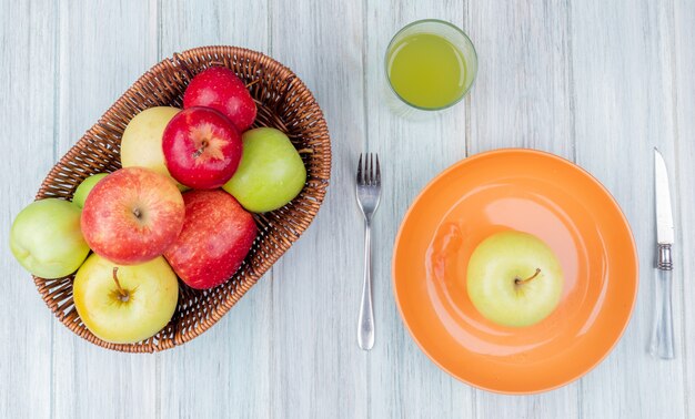 bovenaanzicht van appels in mand en groene in plaat met appelsap mes en vork op houten tafel