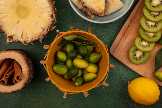 Bovenaanzicht van aromatische kinkans op een emmer met plakjes kiwi op een houten keukenbord met ananas op een kom met kaneelstokjes met citroenen geïsoleerd op een groene achtergrond