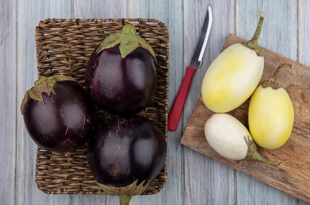 Bovenaanzicht van aubergines in mand plaat met gele en witte op snijplank en mes op houten achtergrond