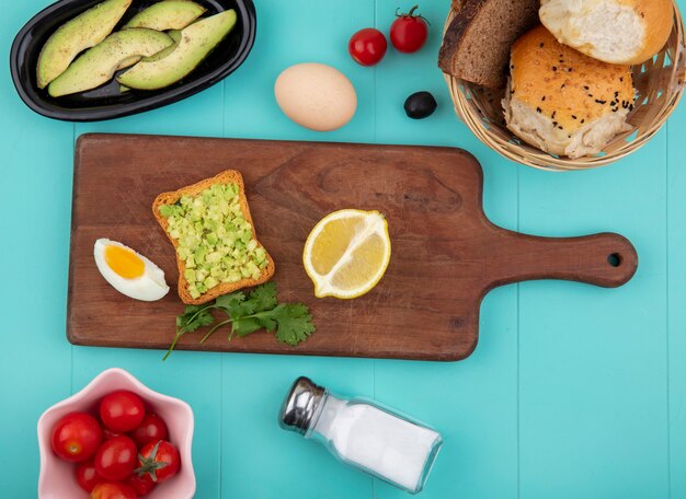 Bovenaanzicht van avocadopulp met geroosterd sneetje brood pn een houten keukenbord met lemonvocado plakjes tomaten op blauw