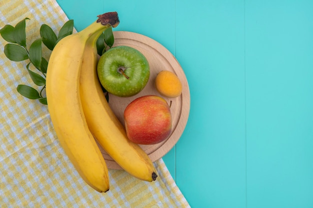 Bovenaanzicht van bananen met appels op een stand op een gele geruite handdoek op een blauwe ondergrond