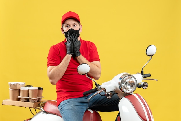 Bovenaanzicht van bezorgde jonge volwassene met rode blouse en muts handschoenen in medisch masker leveren bestelling zittend op scooter op gele achtergrond