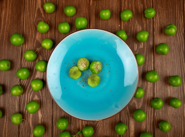 Bovenaanzicht van blauw bord met gesneden groene pruimen bestrooid met gedroogde pepermunt en zure groene pruimen gerangschikt rond op houten tafel
