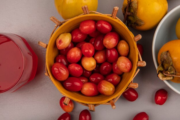 Bovenaanzicht van bleke rode en ovale vorm cornelian fruit op een emmer met kaki fruit op een kom met vers cornelian vruchtensap in een glas op een grijze muur