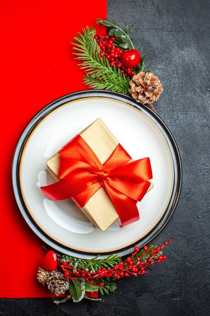 Bovenaanzicht van borden met cadeau erop en dennentakken met decoratie accessoire conifer kegel op een rood servet op een donkere achtergrond